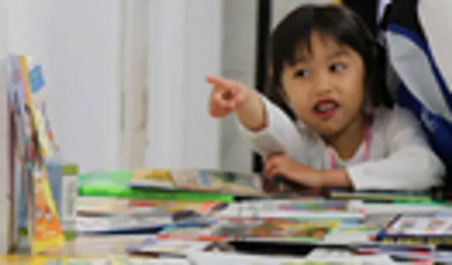 A little girl pointing at the camera while sitting at a table.